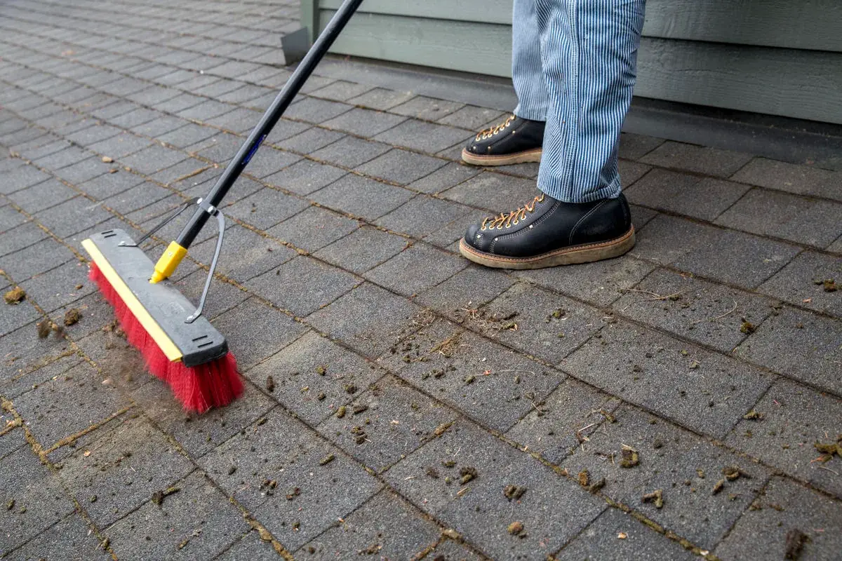 Work boots and a red push broom clearing loosened moss off a shingle roof